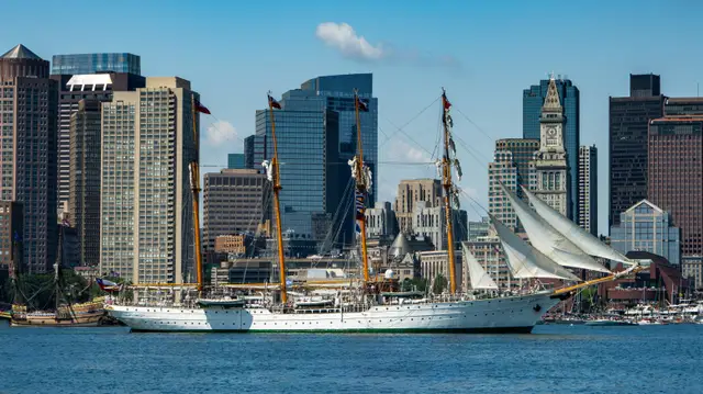 Tall ship under sail on Boston Harbor with the city skyline and Custom House Tower behind.